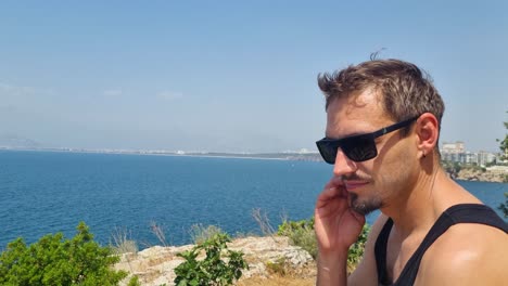 close-up of a man in sunglasses scratching his cheek while sitting on a cliff above the mediterranean sea near antalya, turkey