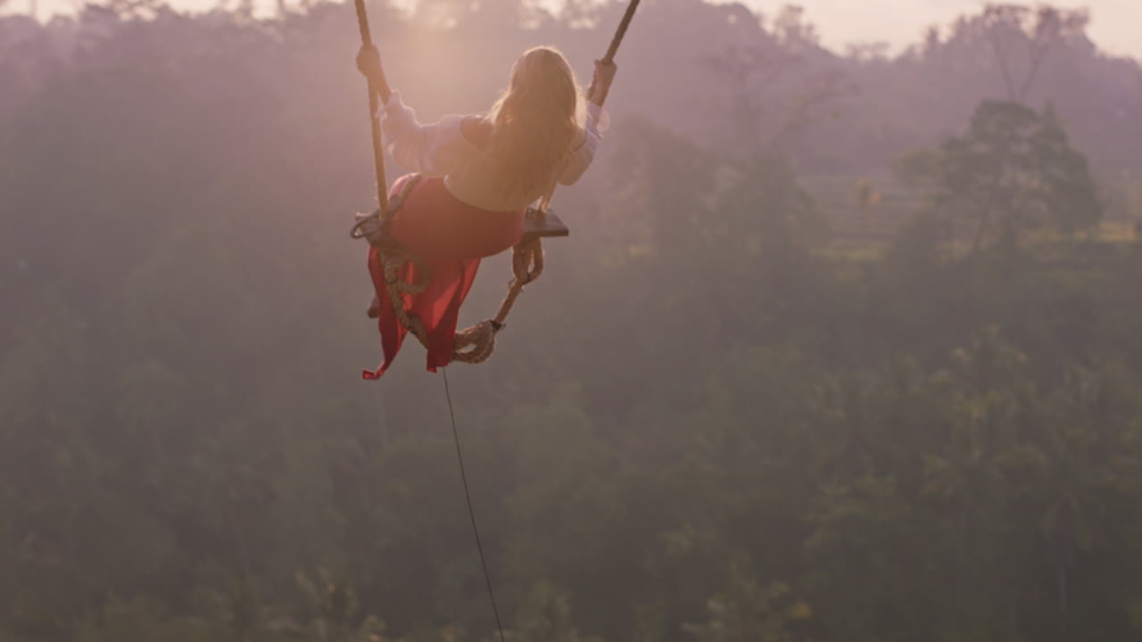 красивая женщина качается над тропическим тропическим лесом на рассвете, сидит на качелях, веселится, наслаждается свободой на экзотическом празднике, образе жизни, замедленном движении.