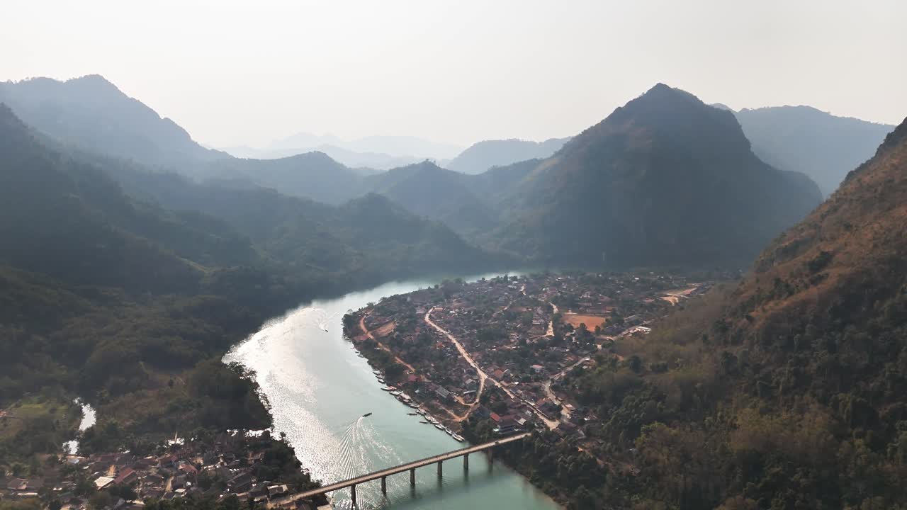 A panoramic view of Nong Khiaw, Laos, with a winding river flanked by towering mountains, a bridge crossing the water, and a small village nestled in the valley under soft afternoon light