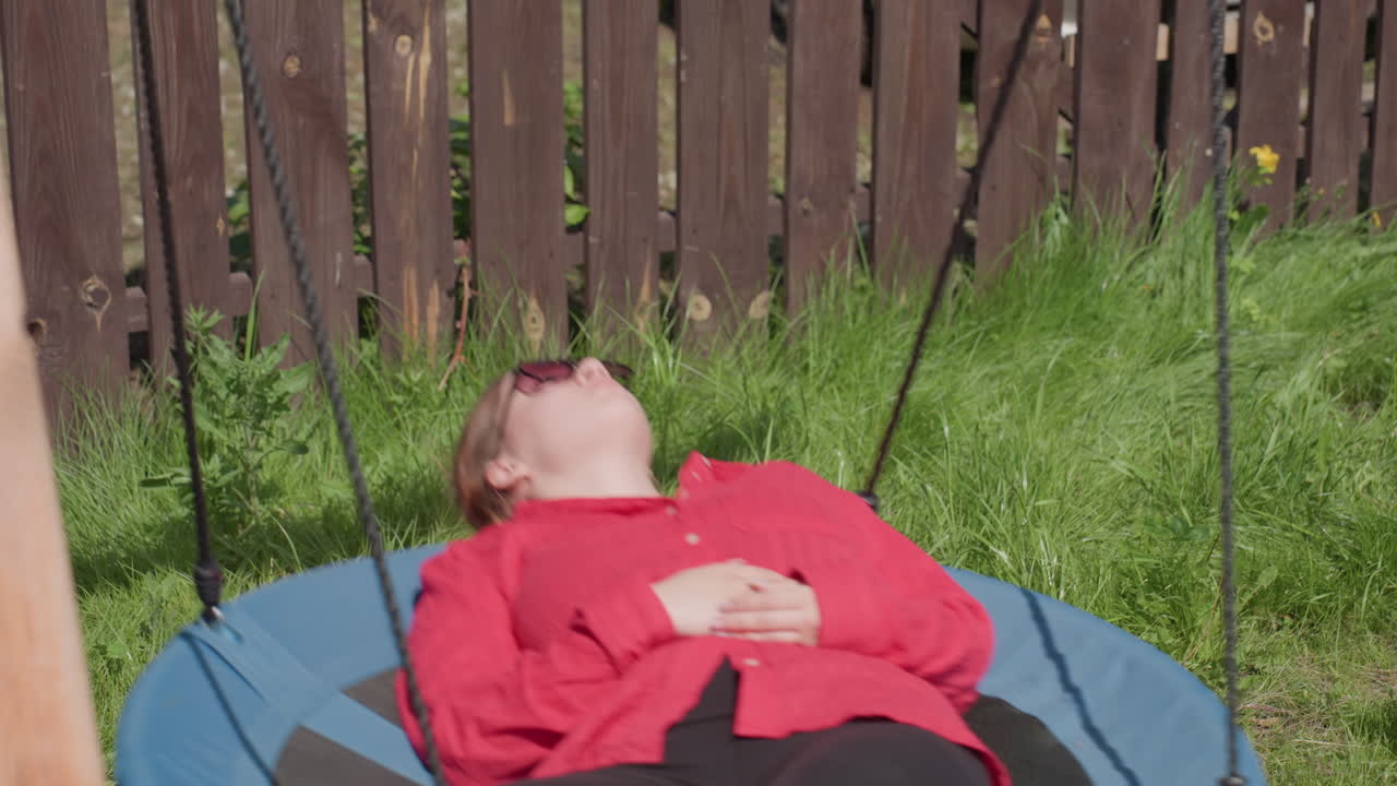 Close up of lady in red shirt laying back on swing with sunglasses under bright sun, sunlight reflects off her face while she rests calmly surrounded by fresh green grass and brown wooden fence