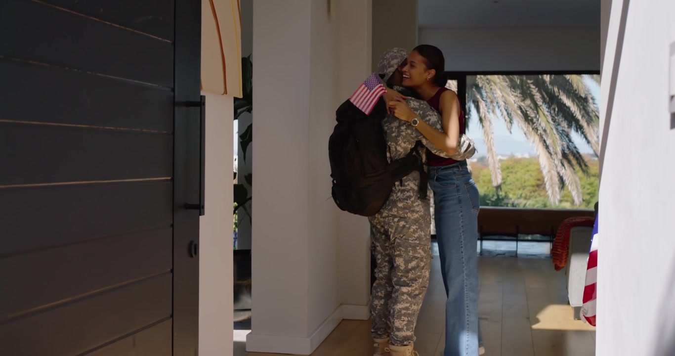 Pareja abrazándose mientras un miembro del servicio entra por la puerta con una mochila de camuflaje, su pareja ondeando una pequeña bandera estadounidense.