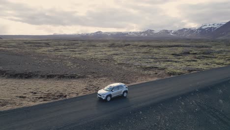 A-car-drives-down-a-road-in-Iceland,-surrounded-by-volcanic-rock-and-moss,-heading-towards-a-mountain-range