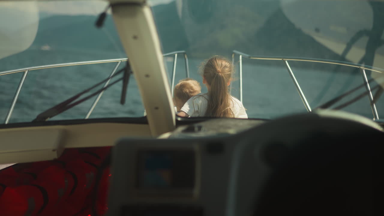niña con el cabello ondeando abrazos hermano pequeño sentado en la cubierta del velero vista desde la cabina. niños encantadores miran en la distancia vela yate en vacaciones de verano