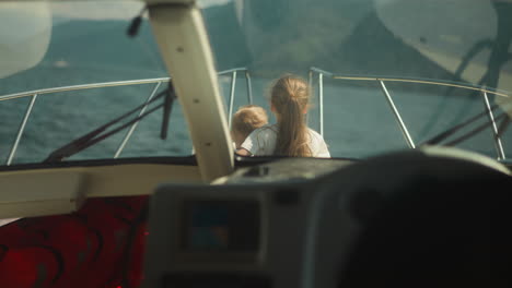 girl with waving hair hugs little brother sitting on sailboat deck view from cockpit. lovely children look in distance sailing yacht on summer vacation