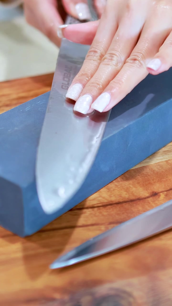 A person sharpens a stainless steel chef’s knife on a blue whetstone atop a wooden cutting board under bright, even kitchen lighting. Close-up, steady camera