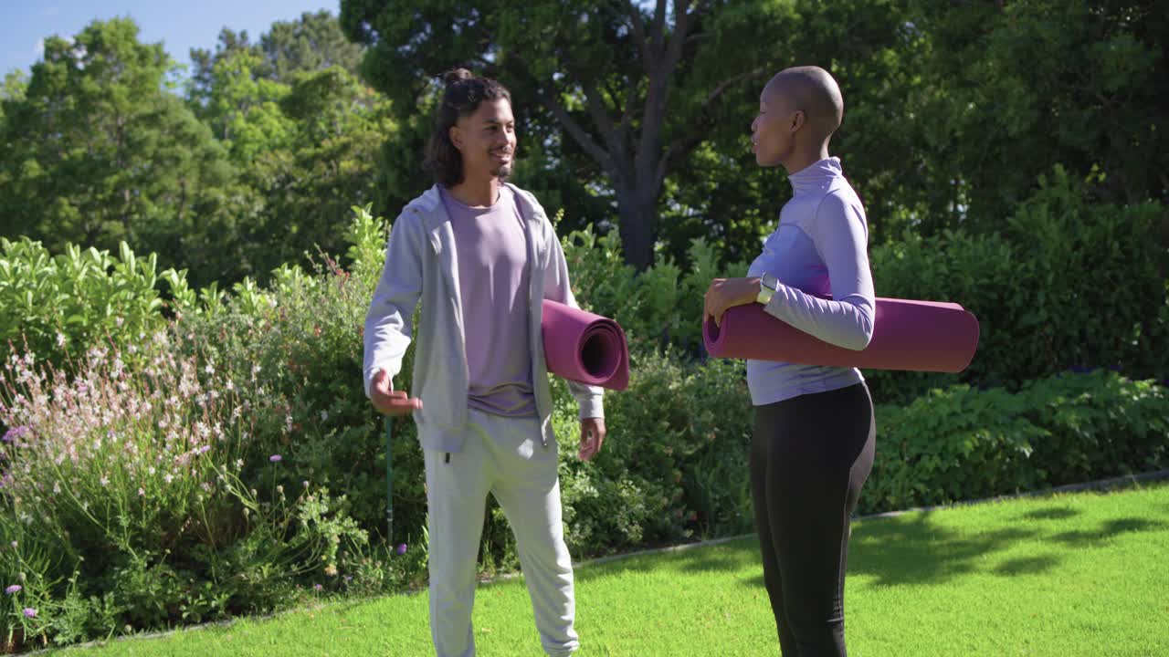 Uomo che cammina mostrando ad amici afroamericani che tengono tappetini, una bottiglia, chiacchierano e si preparano per lo yoga nel parco.