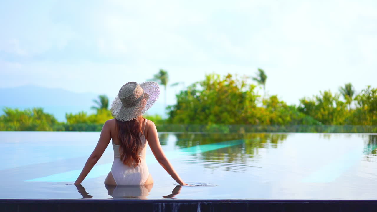 vista trasera de una mujer con sombrero sentada en el borde de la piscina infinita mirando el panorama