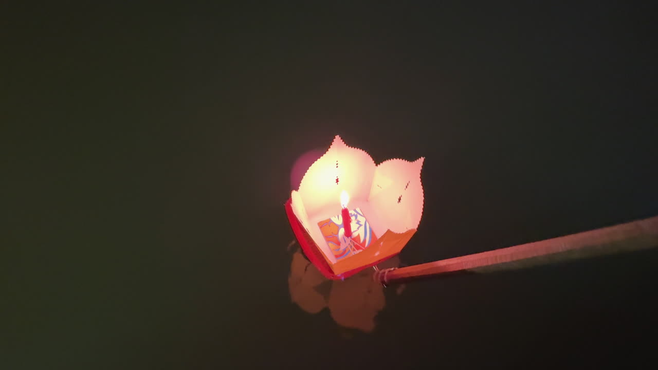 Primo piano di una lanterna di carta luminosa che viene calata da una barca nel fiume Hoài di Hoi An di notte, catturando un tranquillo rituale Tet e il riflesso colorato del festival in Vietnam