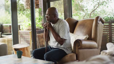 Thoughtful-african-american-senior-man-sitting-in-living-room-leaning-on-walking-stick