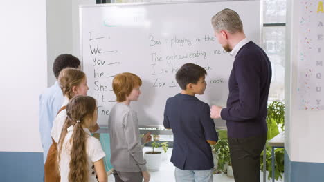 multiethnic students group and teacher standing in front of the blackboard while a ginger student helps an asian classmate to point out the verb to be