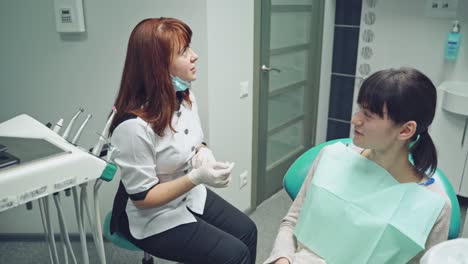 Female doctor explains her patient how to look after the teeth on the background of dental room. Specialist gives five to the patient sitting in the medical chair in the stomatology office.