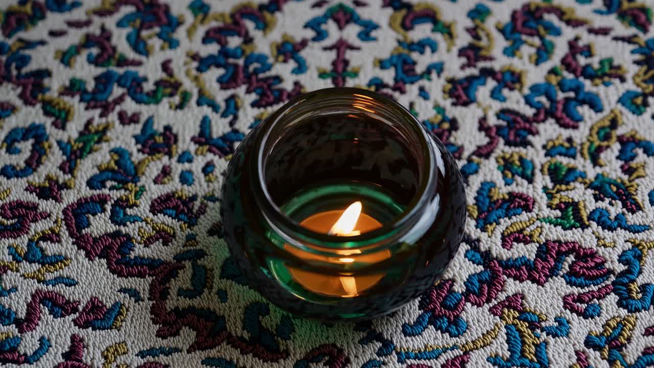 Tealight candle burning inside a round, teal colored glass candle holder, placed on a white tablecloth with colorful embroidered patterns, creating a tranquil and decorative scene