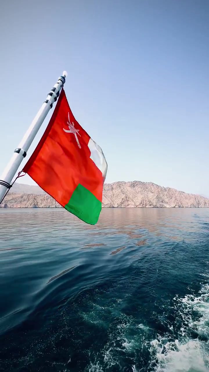 National flag waves from moving boat above calm sea waters with rugged mountain coastline in background, showcasing maritime travel in Oman