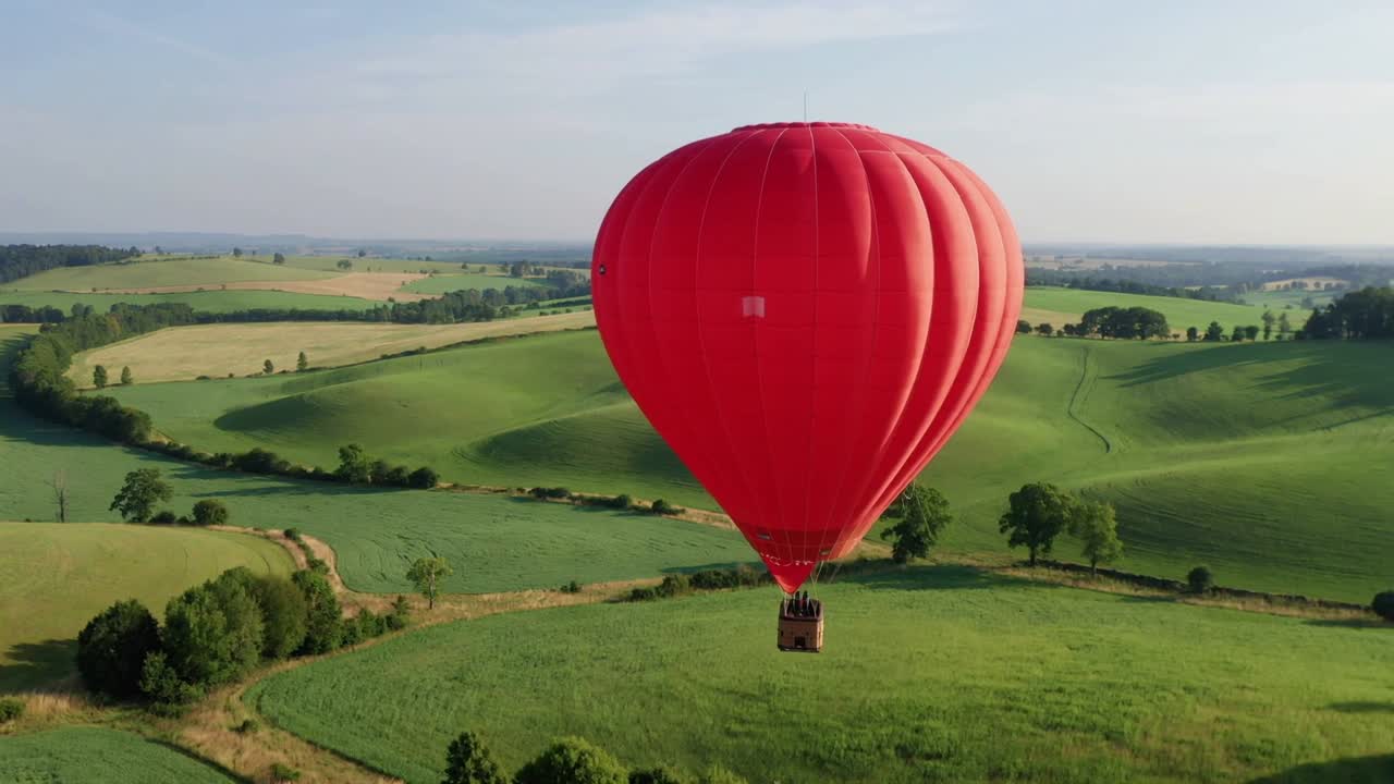 Câmera de rastreamento capturando balão de ar quente vermelho flutuando sobre terras agrícolas com passageiros em cesta de vime.