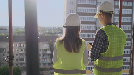 Back-turned-architect-and-technician-at-construction-site-reviewing-office-blueprints