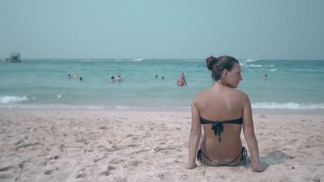 brunette-in-black-bikini-sits-on-yellow-sand-ocean-beach