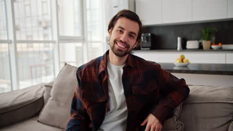 Portrait-of-a-happy-brunette-guy-with-stubble-in-a-red-blue-checkered-shirt-who-sits-on-a-modern-brown-sofa-in-a-bright-apartment-with-large-windows