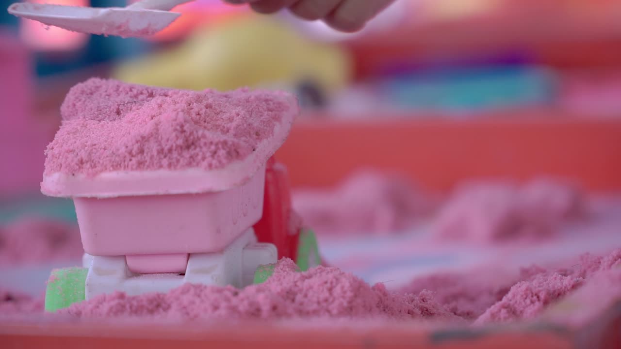 Child's hands are playing with pink sand using a toy shovel