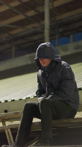 un hombre vestido con un traje negro y una chaqueta se sienta solo en un estadio tranquilo con las manos juntas entre las piernas y una pelota cerca de él ligeramente movida por el viento