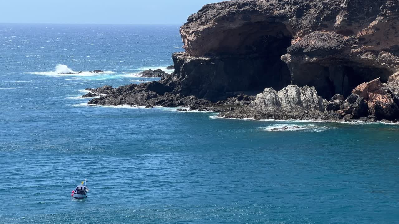A small fishing boat sails near the rugged volcanic cliffs and sea caves of Ajuy, Fuerteventura, with waves crashing on the rocks. Canary Islands, Spain