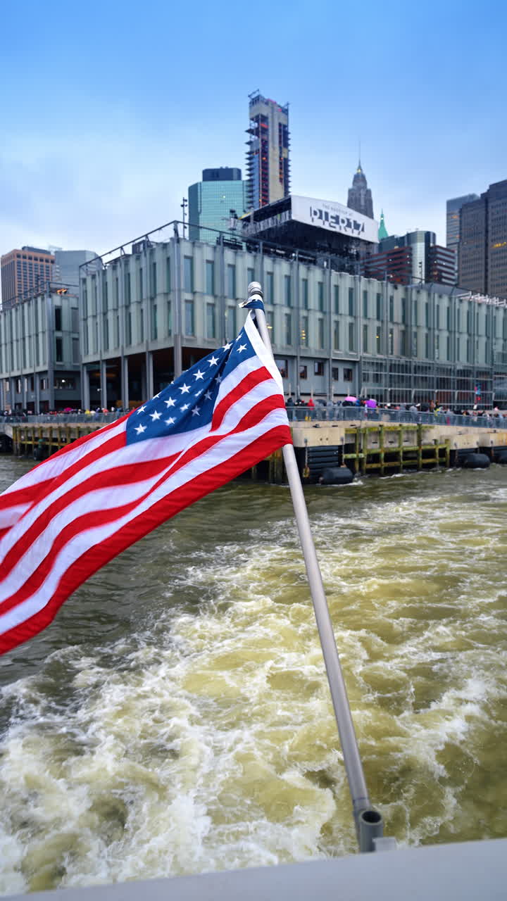 Bandiera americana che sventola su una barca che lascia il molo di New York. Bandiera degli Stati Uniti in primo piano con il Molo 17 e gli edifici della città sullo sfondo.