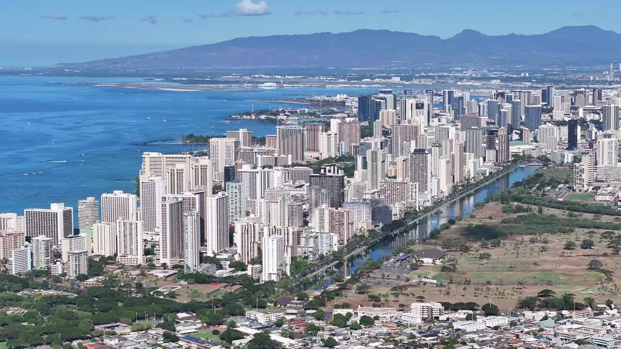 Aerial drone footage overlooking the Honolulu skyline on Oahu, Hawaii, showcasing high-rise hotels, tropical coastline, blue ocean waters, and the scenic cityscape of Waikiki under sunny skies