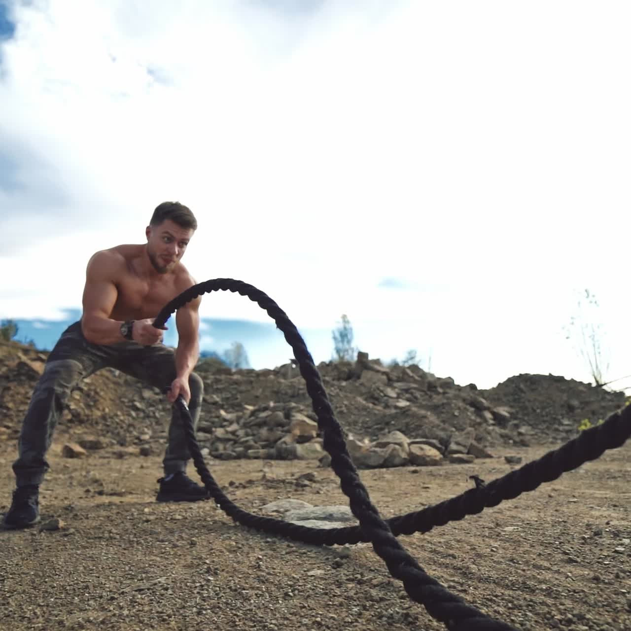 Muscular athlete doing waves with battle ropes in nature. Professional sportsman exercises with cables outdoors. Healthy lifestyle.