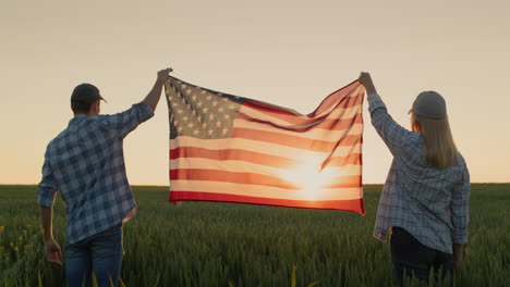 two happy farmers raise the us flag over a field of wheat as the sun sets. july 4th - independence day usa concept