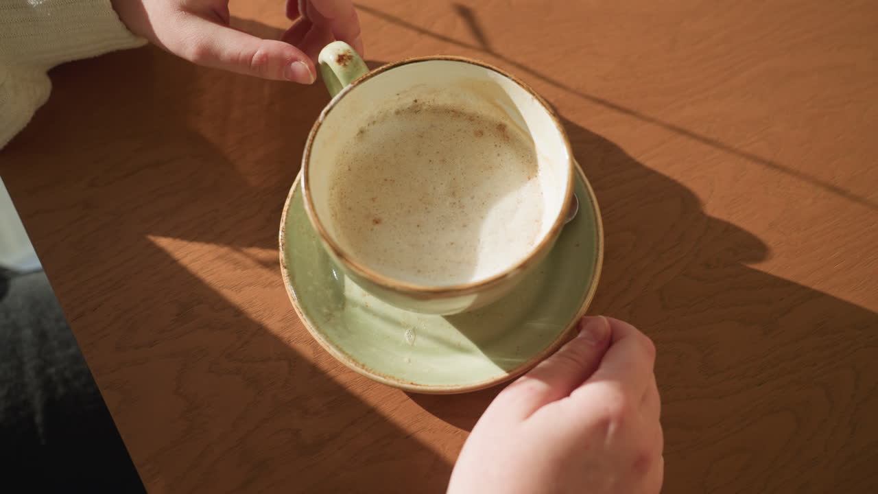 Close up of woman hands gently rotating ceramic coffee cup on wooden table, foam visible inside cup, soft daylight casting shadows across surface