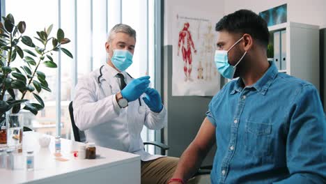 Portrait-Of-Handsome-Middle-Aged-Male-Professional-Physician-Make-Covid-19-Vaccine-Injection-To-Young-Hindu-Man-Patient-In-Medical-Mask-Sitting-In-Hospital-Room,-Coronavirus-Vaccination-In-Quarantine