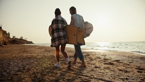 Rückansicht-Eines-Blonden-Mädchens-Mit-Grünem-Hut-Und-Kariertem-Hemd-Und-Ihres-Brünetten-Freundes-In-Weißem-T-Shirt,-Die-Am-Sandstrand-Entlang-Des-Meeres-Spazieren-Und-Morgens-Bei-Sonnenaufgang-Surfbretter-Tragen