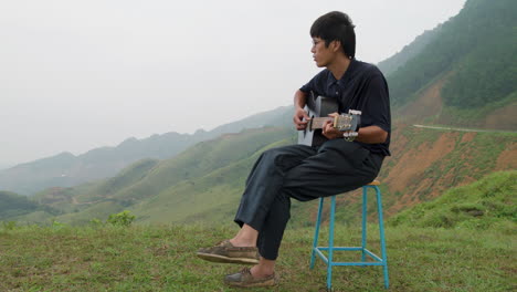 talented asian musician man playing the guitar sitting on a stool in middle of green mountains