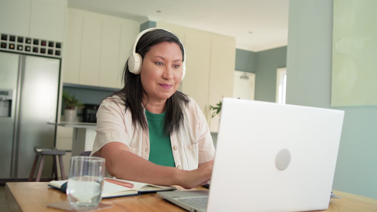Woman approaching kitchen table, grabbing papers and phone, wearing headphones, joining laptop call