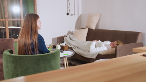 therapist seated on green armchair facing guest lying on reclining cushion on brown sofa inside contemplative room, warm wooden accents, clock and tissue box on table between them now
