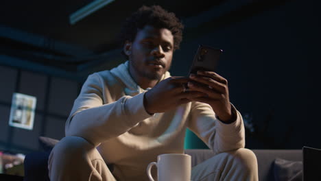 smiling african american man enjoying a moment of fun typing messages on a mobile phone, sitting in a cozy living room. adult in a relaxed atmosphere reflects work from home flexibility. camera a.