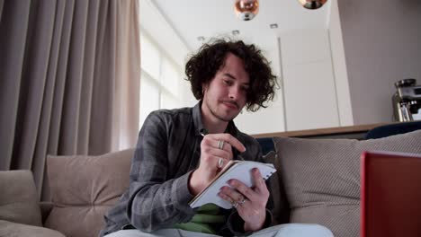 Focused-brunette-guy-with-curly-hair-and-mustache-in-a-gray-checkered-shirt-takes-notes-and-writes-down-important-points-while-watching-an-educational-video-on-a-red-laptop-while-sitting-on-the-couch-at-home