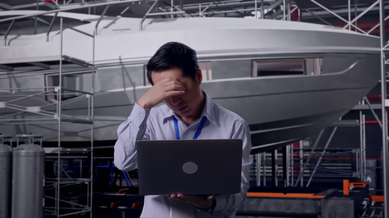 Asian Male Professional Worker Standing With His Laptop with Yacht Construction in Modern Shipyard, He Is Nodding His Head With Dissapionted