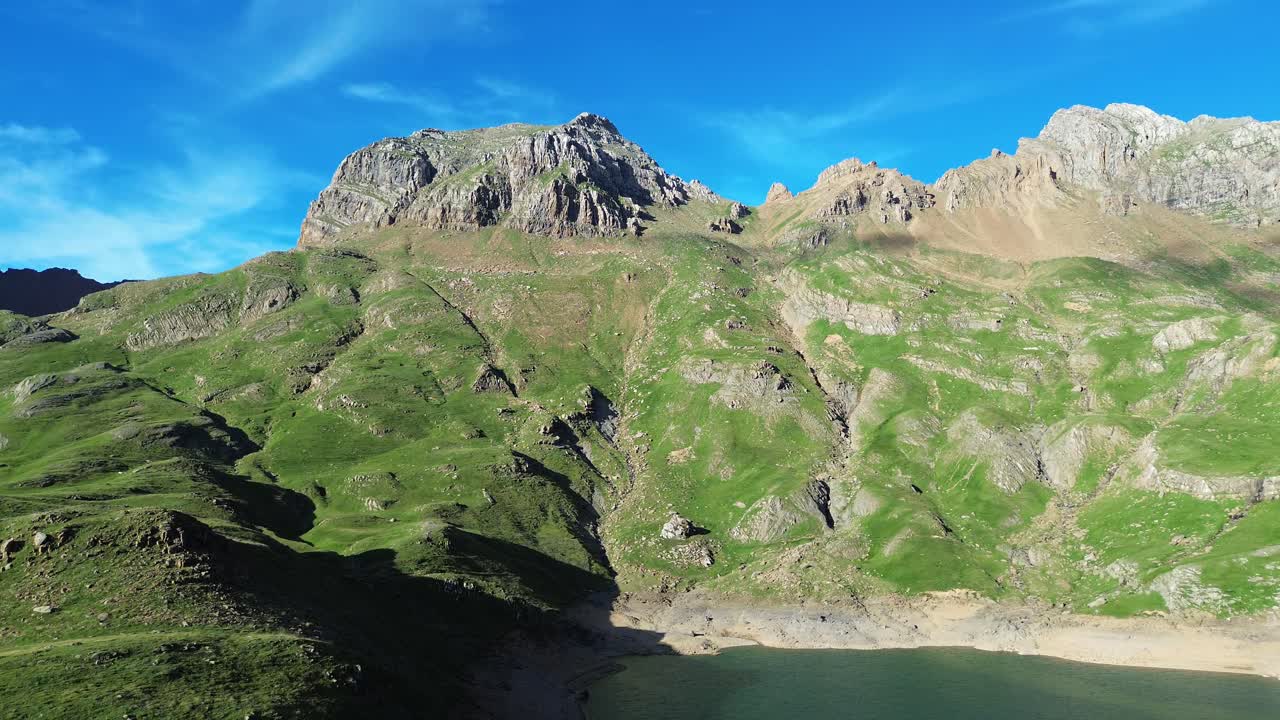 Aerial footage of Valle de Ip near Canfranc, Spain, showcasing majestic green mountains and a tranquil lake under a vibrant blue sky. Captures nature's serenity and rugged beauty