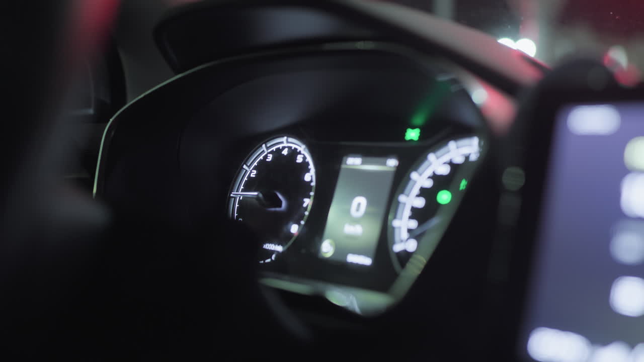 Close up of car dashboard at night showing glowing speedometer and tachometer, dim ambient reflections on windshield, blurred lights from outside street visible in background
