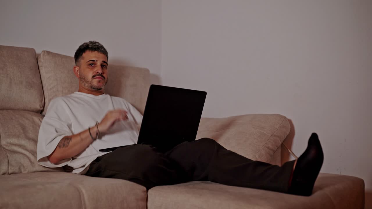 Young man reclining on sofa with legs extended while working on laptop at home