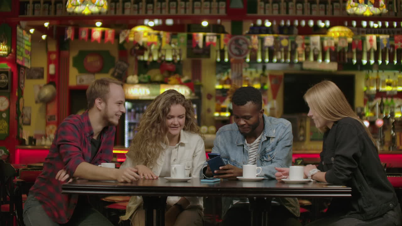 Portrait of cheerful young friends looking at smart phone while sitting in cafe. Mixed race people sitting at a table in restaurant using mobile phone