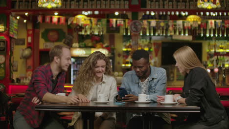 retrato de jóvenes amigos alegres mirando un teléfono inteligente mientras están sentados en un café. personas de raza mixta sentadas en una mesa en un restaurante usando un teléfono móvil