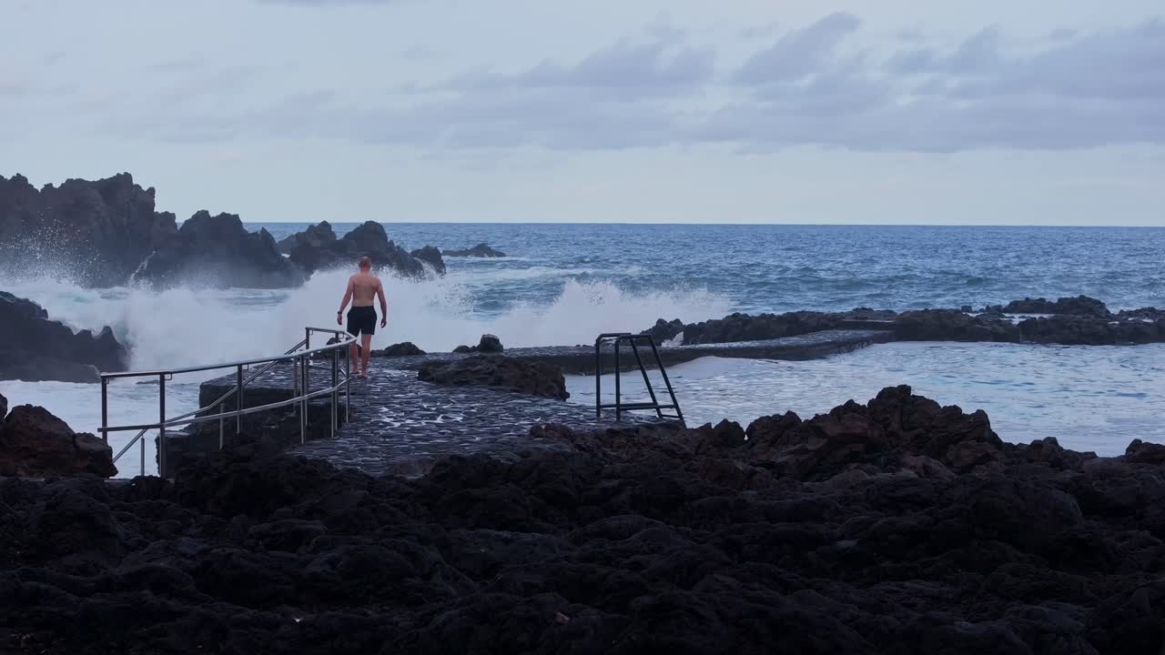 Ocean swells crash near volcanic coast as man walks calmly on stone path
