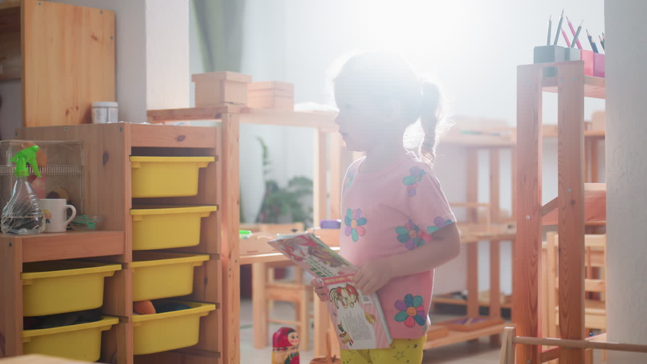 Seorang gadis kecil di taman kanak-kanak berdiri dengan buku di tangan tampak terganggu saat cahaya matahari alami masuk melalui jendela, furnitur kayu, tempat penyimpanan kuning, tanaman, dan latar belakang ruang kelas yang nyaman