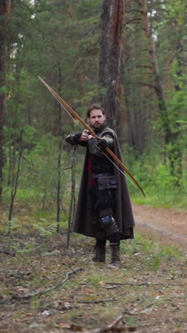 warrior with cape and armour shoots bow looking around on road in green forest slow motion. archer in medieval costume. historical reconstruction