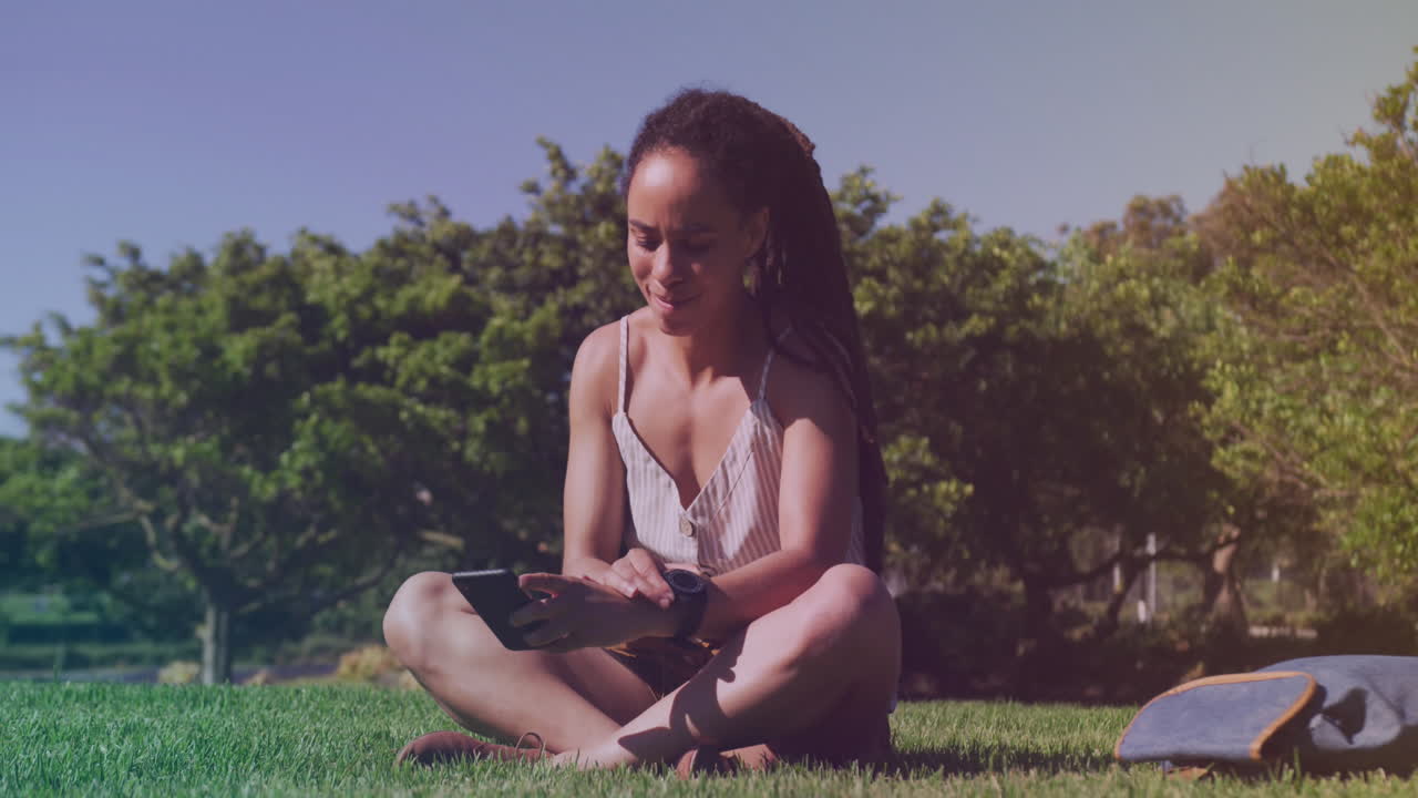 Spots of light against african american woman using smartphone sitting on the grass