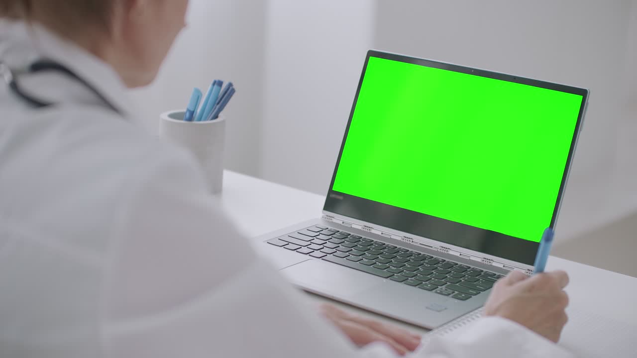 la doctora está aprendiendo en línea, mirando la pantalla verde de la computadora portátil para la tecnología de llave cromática, escribiendo notas en papel y asintiendo con la cabeza