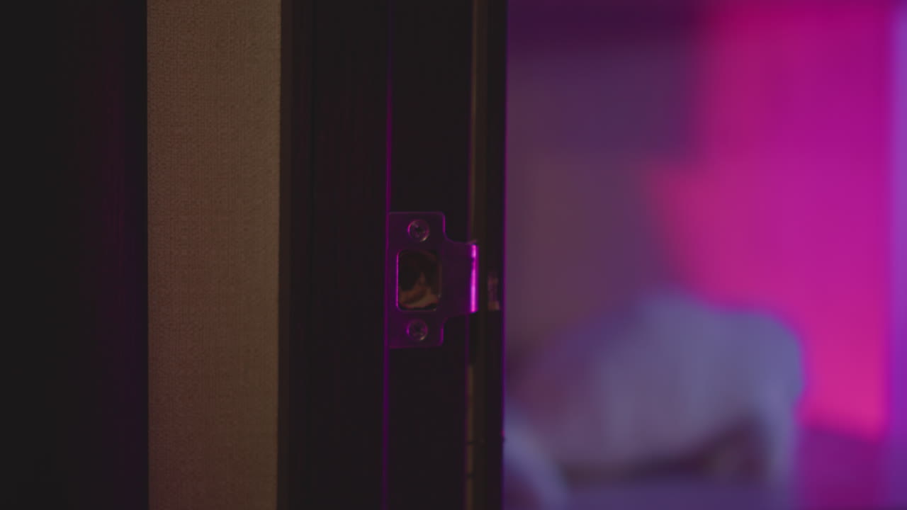 Close up hand in fluffy sweater gripping metal knob and turning door open to enter neon lit studio bathed in pink purple hue as silhouette steps through threshold under moody ambient glow