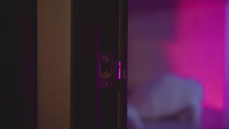 close up hand in fluffy sweater gripping metal knob and turning door open to enter neon lit studio bathed in pink purple hue as silhouette steps through threshold under moody ambient glow