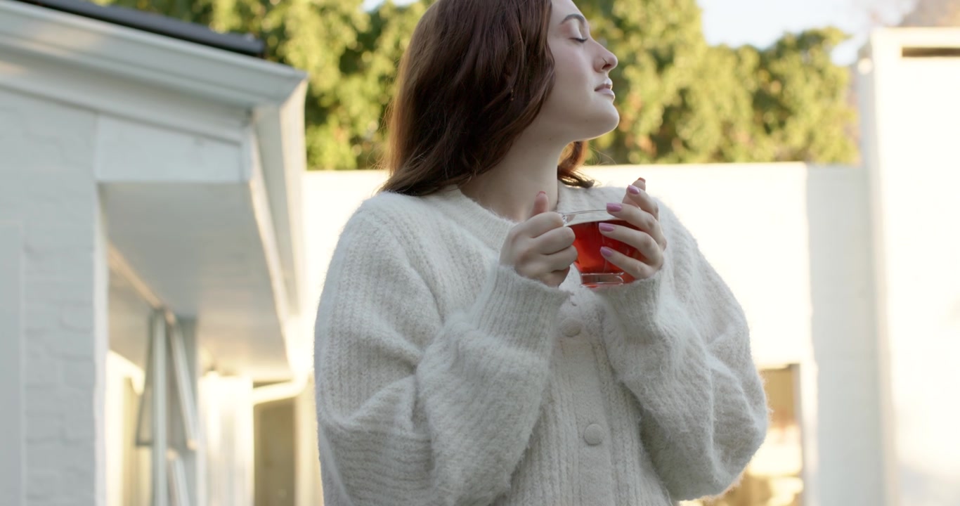Donna in piedi sul balcone con un cardigan bianco, che tiene una tazza rossa mentre la telecamera panoramica, guardando oltre la ringhiera